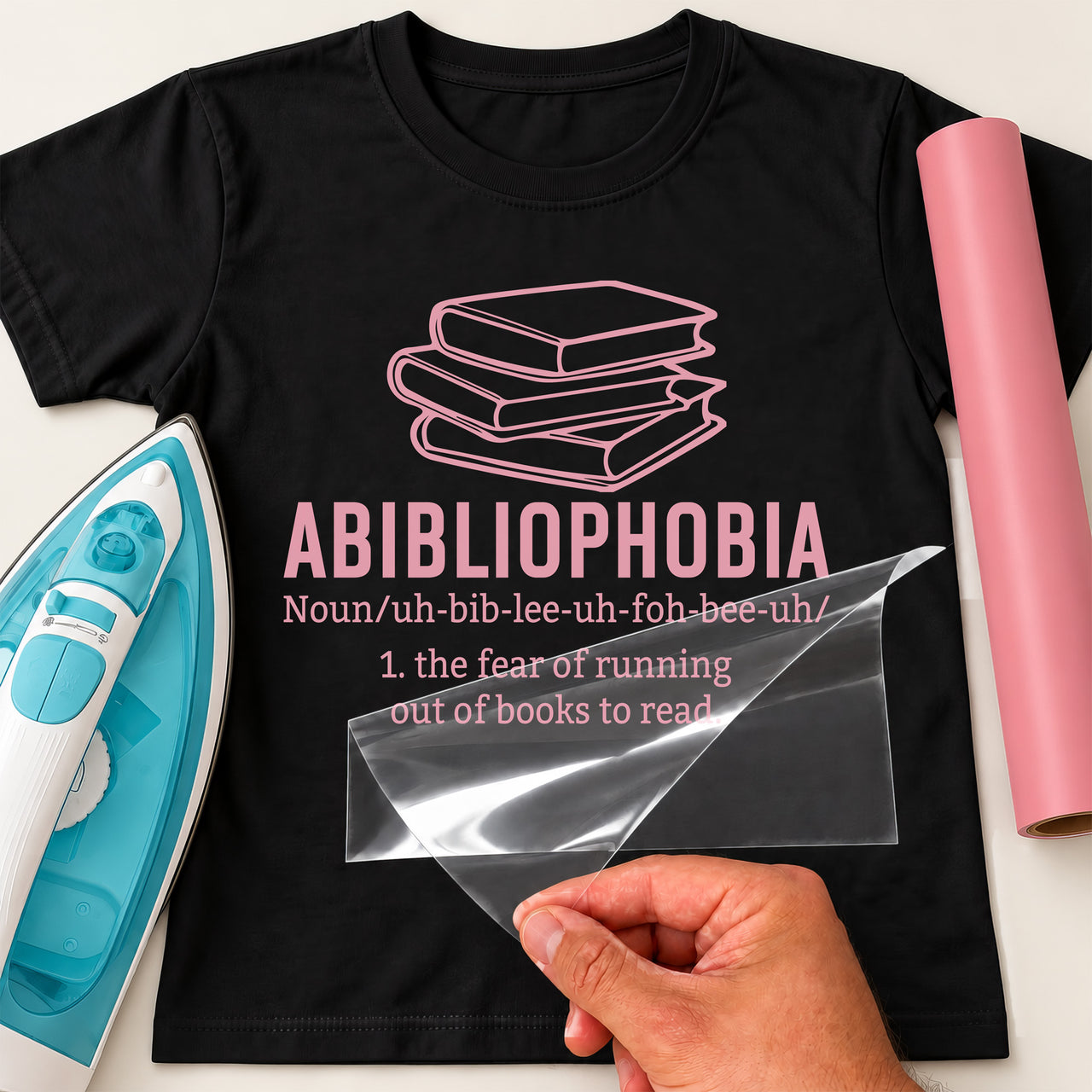 Black T-shirt with pink iron-on vinyl: stack of books and 'ABIBLIOPHOBIA' definition, iron and pink vinyl roll beside