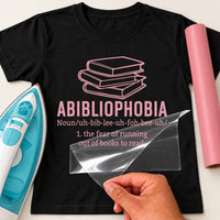 Black T-shirt with pink iron-on vinyl: stack of books and 'ABIBLIOPHOBIA' definition, iron and pink vinyl roll beside