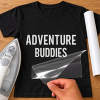 Black T-shirt with white 'ADVENTURE BUDDIES' iron-on vinyl transfer, carrier sheet being peeled off, iron beside