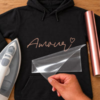 Black hoodie with rose gold script 'Amour' ending in a small heart, carrier film being peeled off - iron-on vinyl transfer