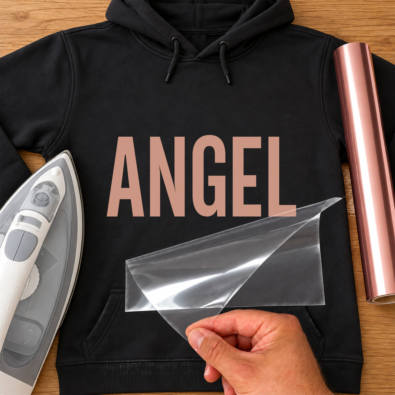 Black hoodie with rose gold 'ANGEL' iron-on vinyl transfer in bold capitals, carrier sheet being peeled off, iron