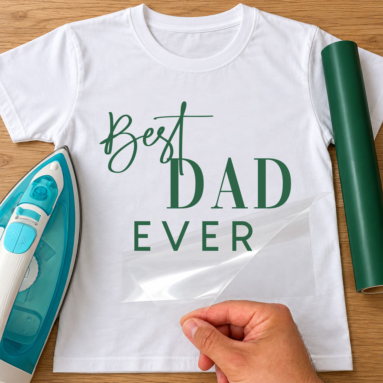 White T-shirt with green script and serif dad lettering, film peeled back beside a teal iron