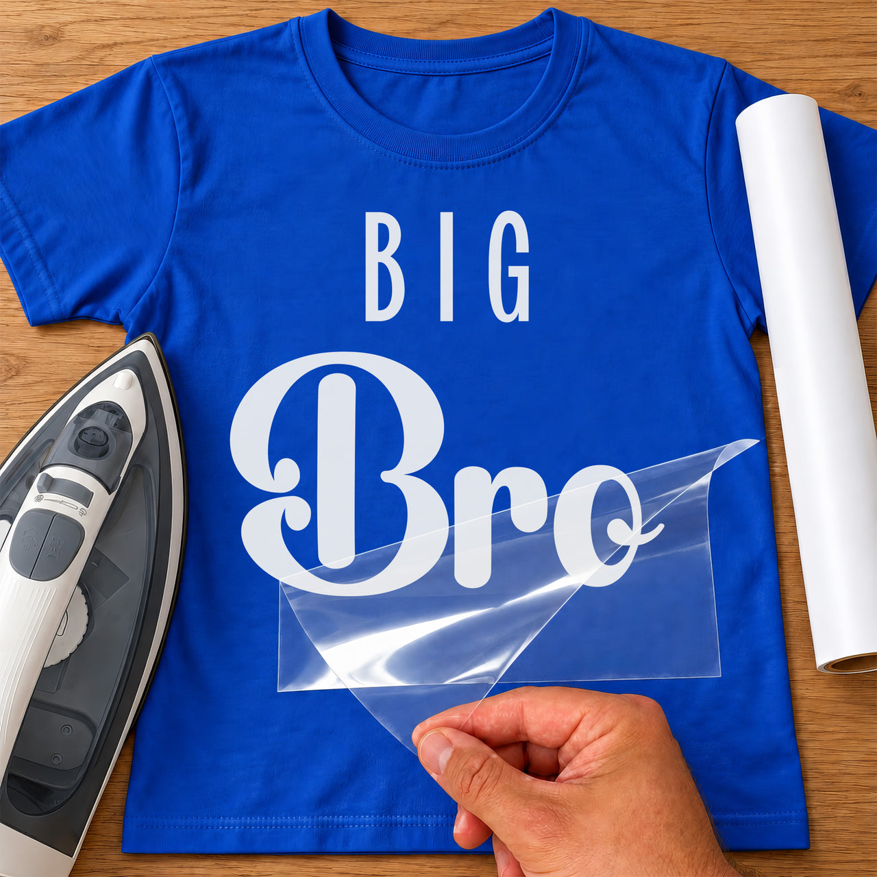 Royal blue T-shirt with white iron-on vinyl reading 'BIG Bro' being peeled, iron and white vinyl roll beside