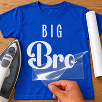 Royal blue T-shirt with white iron-on vinyl reading 'BIG Bro' being peeled, iron and white vinyl roll beside