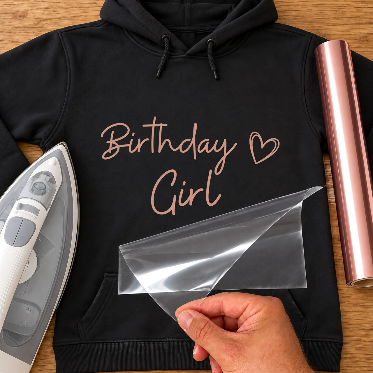 Birthday Girl iron-on transfer: rose gold Birthday Girl script with a heart on a black hoodie, film being peeled