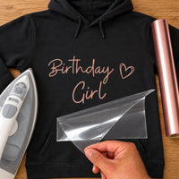 Birthday Girl iron-on transfer: rose gold Birthday Girl script with a heart on a black hoodie, film being peeled