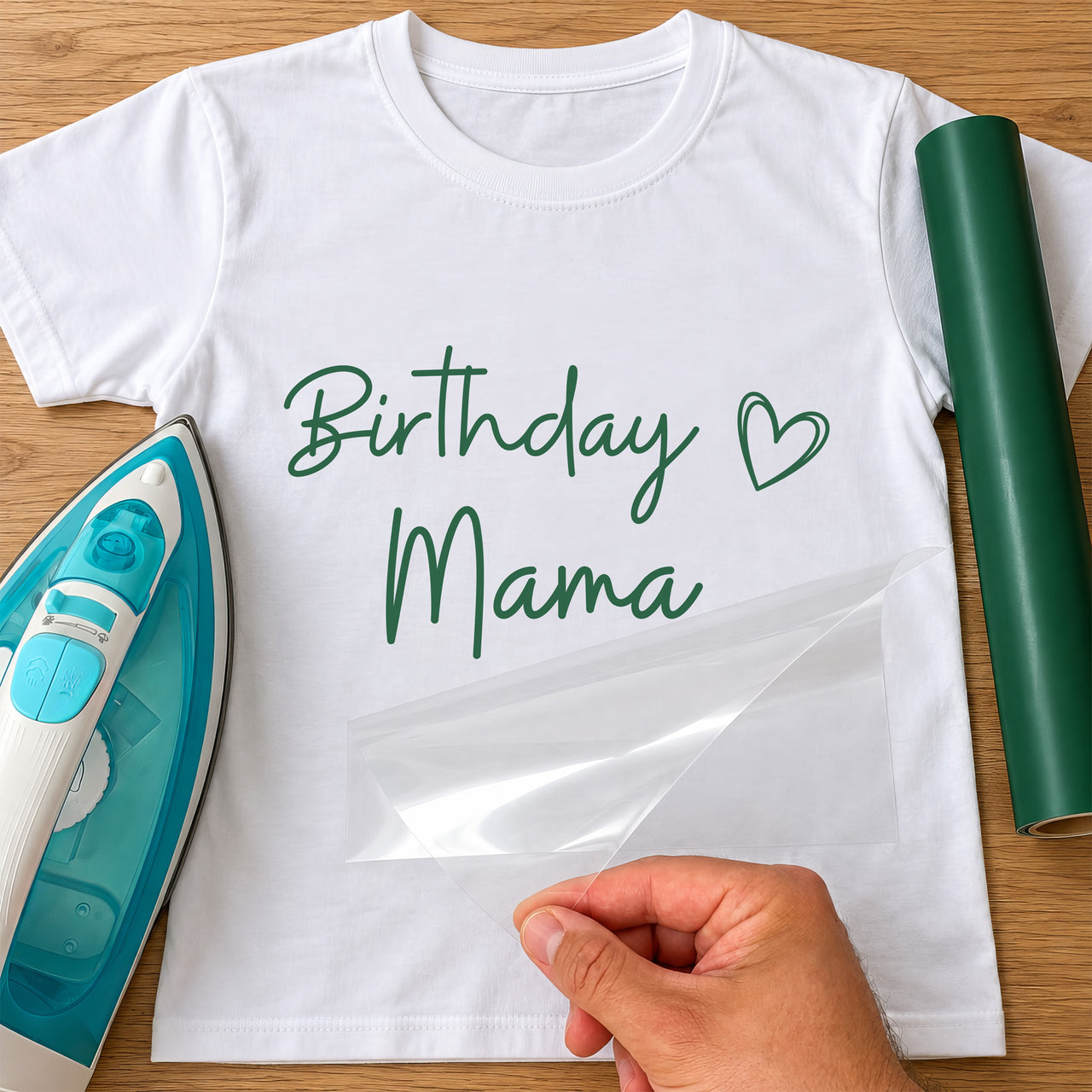 Green 'Birthday Mama' handwritten iron-on transfer with heart on a white T-shirt beside a vinyl roll