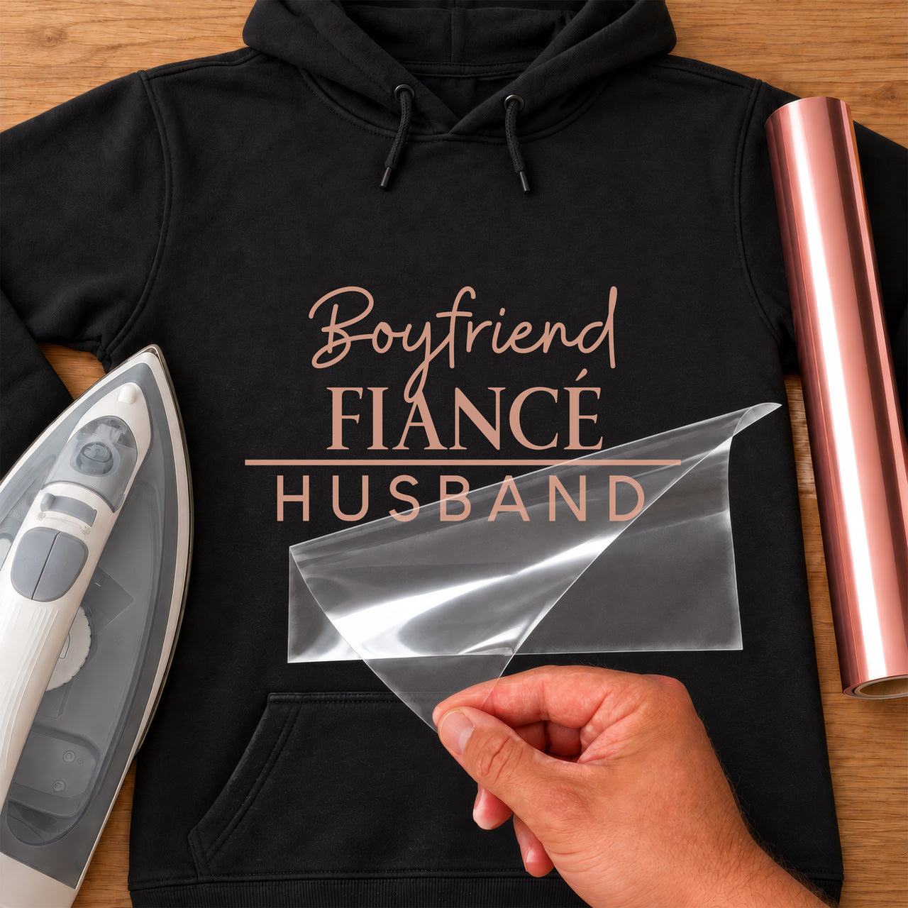 Black hoodie with rose gold iron-on vinyl reading 'Boyfriend FIANCÉ HUSBAND', iron and rose gold vinyl roll beside