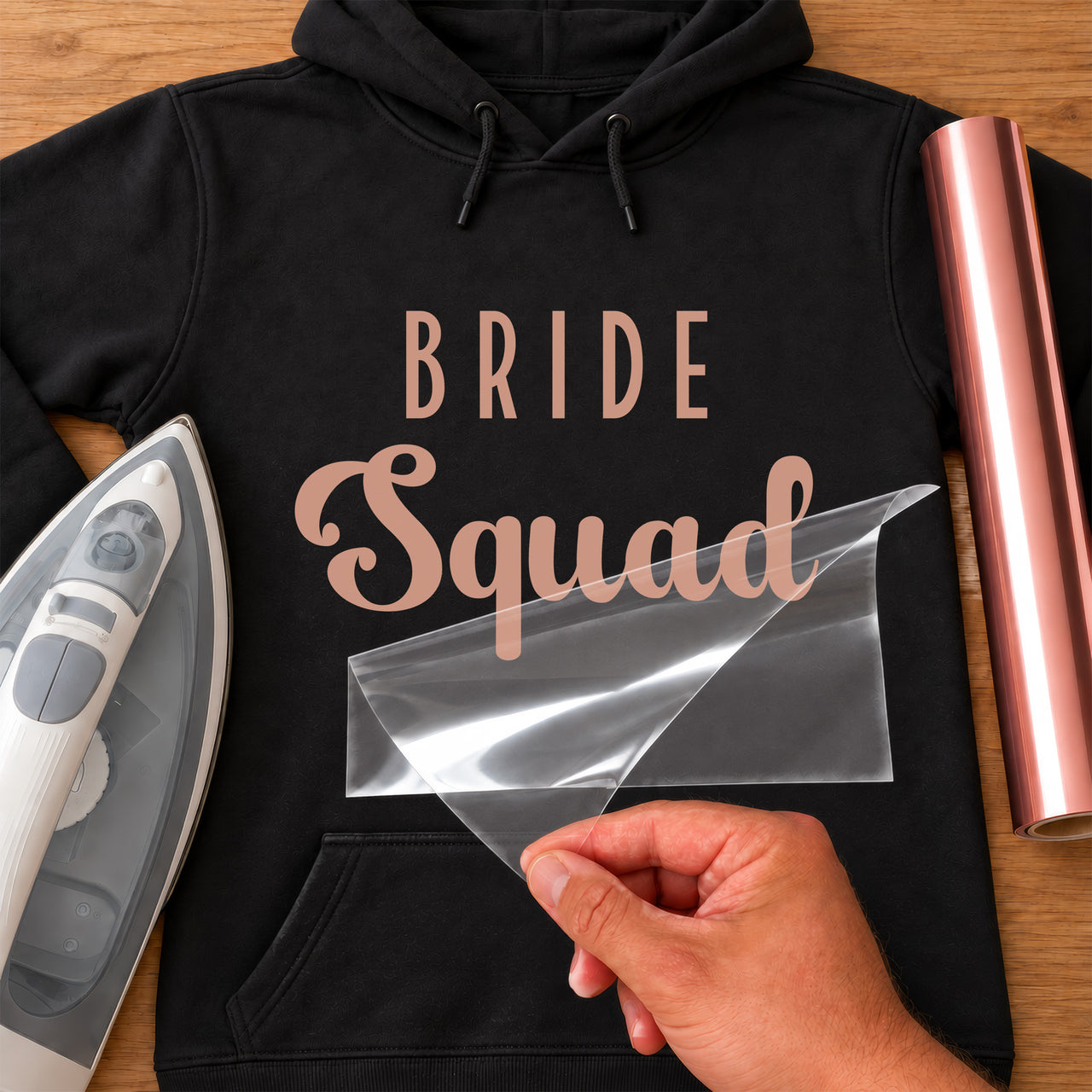 Rose gold "Bride Squad" iron-on vinyl on a black hoodie with iron, vinyl roll and peeled carrier sheet