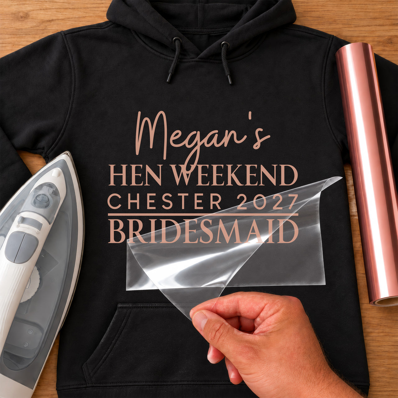 Rose gold vinyl on a black hoodie: 'Megan's Hen Weekend Chester 2027 Bridesmaid', mockup with iron and film