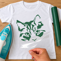 White T-shirt with a green stencil-style cat face, iron and green vinyl roll beside, iron-on vinyl transfer