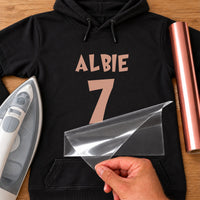 Black hoodie mockup with rose gold iron-on name 'ALBIE' and a large number 7, carrier sheet peeling off