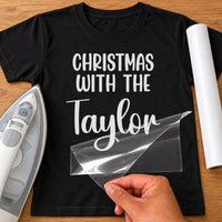 Christmas With The Family iron-on transfer: white CHRISTMAS WITH THE Taylor on a black T-shirt, film being peeled