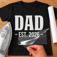Black T-shirt with white 'DAD' and 'EST. 2026' iron-on transfer in bold block letters, iron and white vinyl roll
