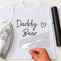 Mockup of white T-shirt printed with handwritten 'Daddy Bear' and outline heart, iron and black vinyl beside it