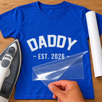 Blue T-shirt with white 'DADDY ~ EST. 2026 ~' lettering, carrier film being peeled off - iron-on vinyl transfer