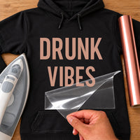 Black hoodie with 'DRUNK VIBES' in rose gold vinyl, iron and vinyl roll beside, iron-on transfer
