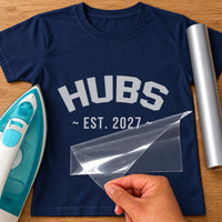 Navy T-shirt with grey iron-on vinyl reading 'HUBS ~ EST. 2027 ~' being peeled, iron and grey vinyl roll beside