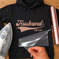 Husband Since iron-on transfer: rose gold Husband script with swoosh and SINCE 2015 on a black hoodie, film being peeled