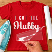 Red T-shirt with white 'I GOT THE Hubby' iron-on vinyl transfer, carrier sheet being peeled off, iron and vinyl roll