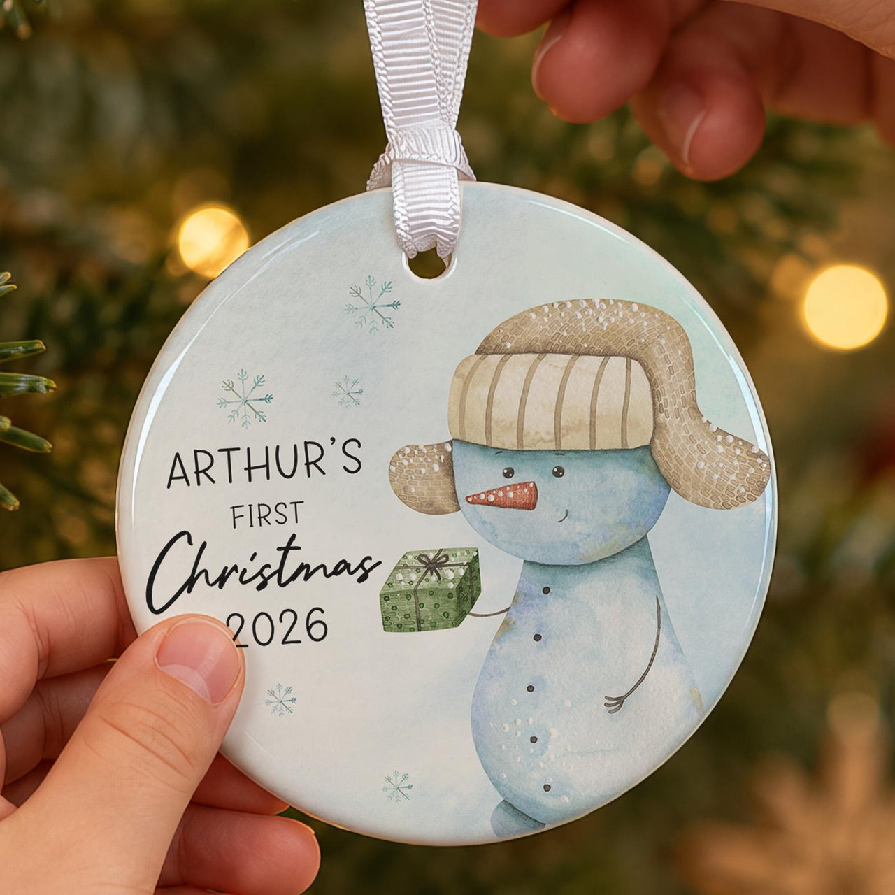 First Christmas bauble: pale blue ornament with a snowman in a fur hat holding a gift and ARTHUR'S First Christmas 2026