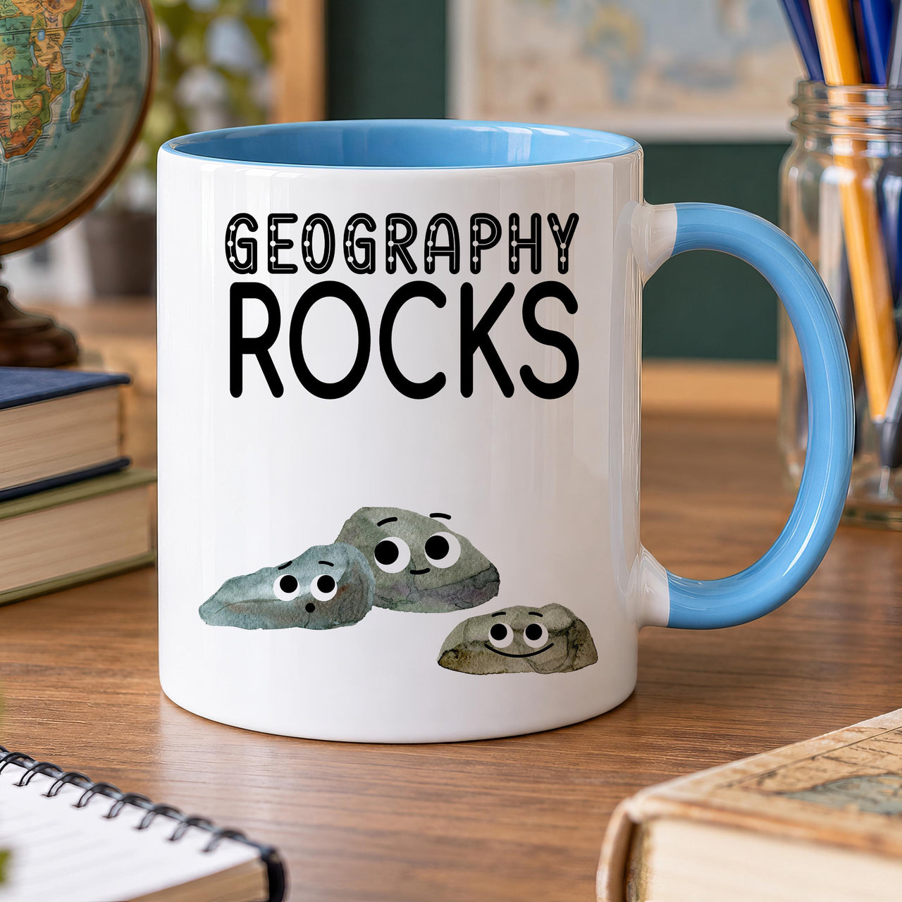 White mug with a light blue handle printed Geography Rocks above three cartoon pebbles