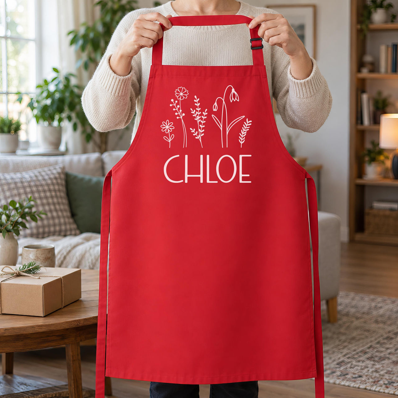 Red apron with white wildflowers and 'CHLOE', held up by the neck strap in a living room - personalised apron