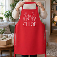 Red apron with white wildflowers and 'CHLOE', held up by the neck strap in a living room - personalised apron