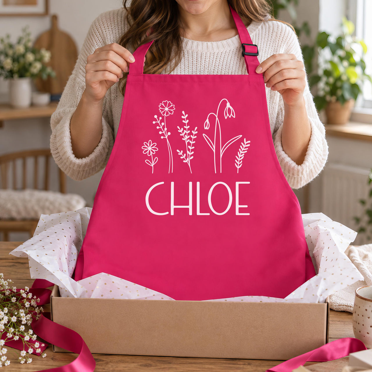 Pink apron with white wildflowers and 'CHLOE', lifted from a gift box with tissue paper - personalised apron