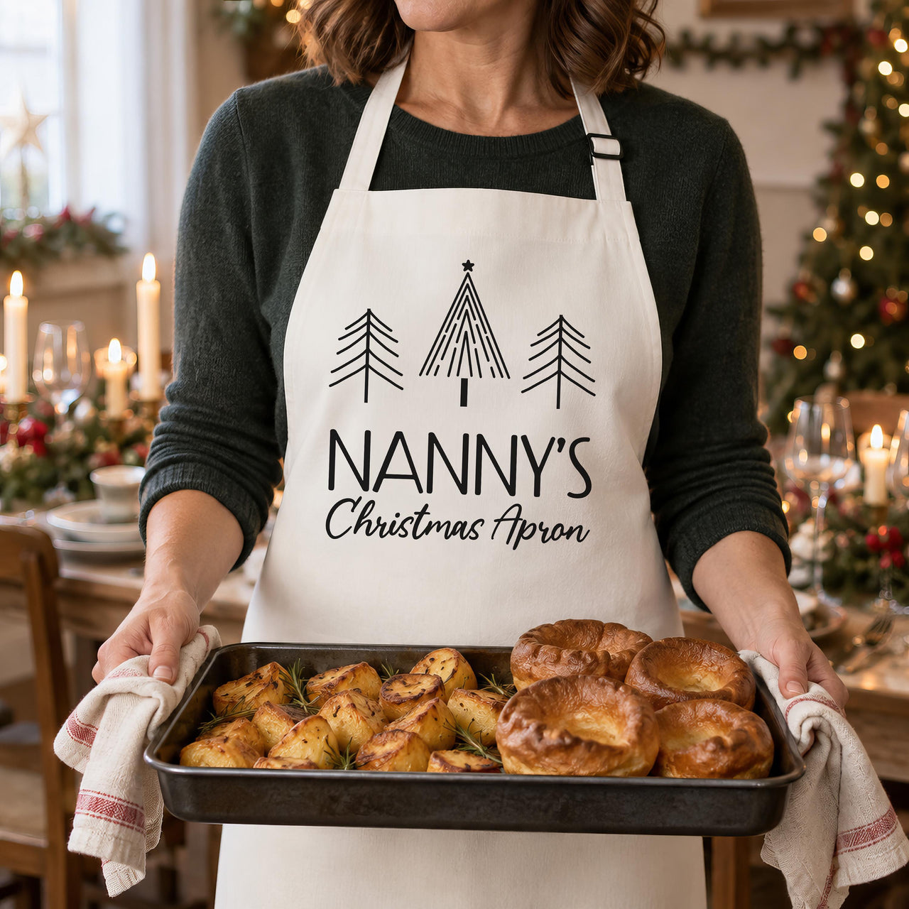White Nanny's Christmas Apron with black tree print, worn while holding a tray of pastries