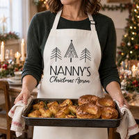 White Nanny's Christmas Apron with black tree print, worn while holding a tray of pastries