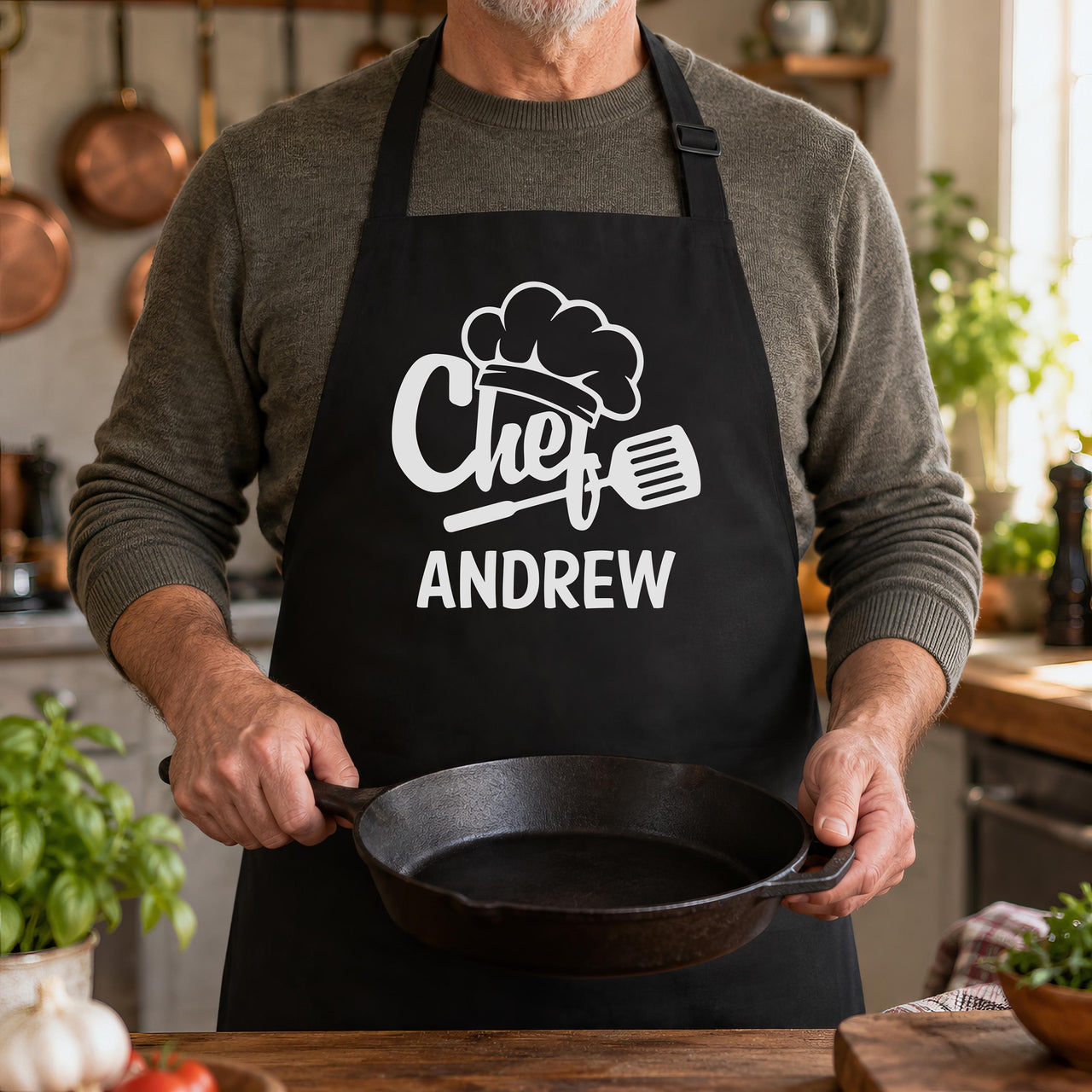 Black apron printed with a chef hat, "Chef" and "ANDREW", worn while holding a cast iron pan - personalised apron
