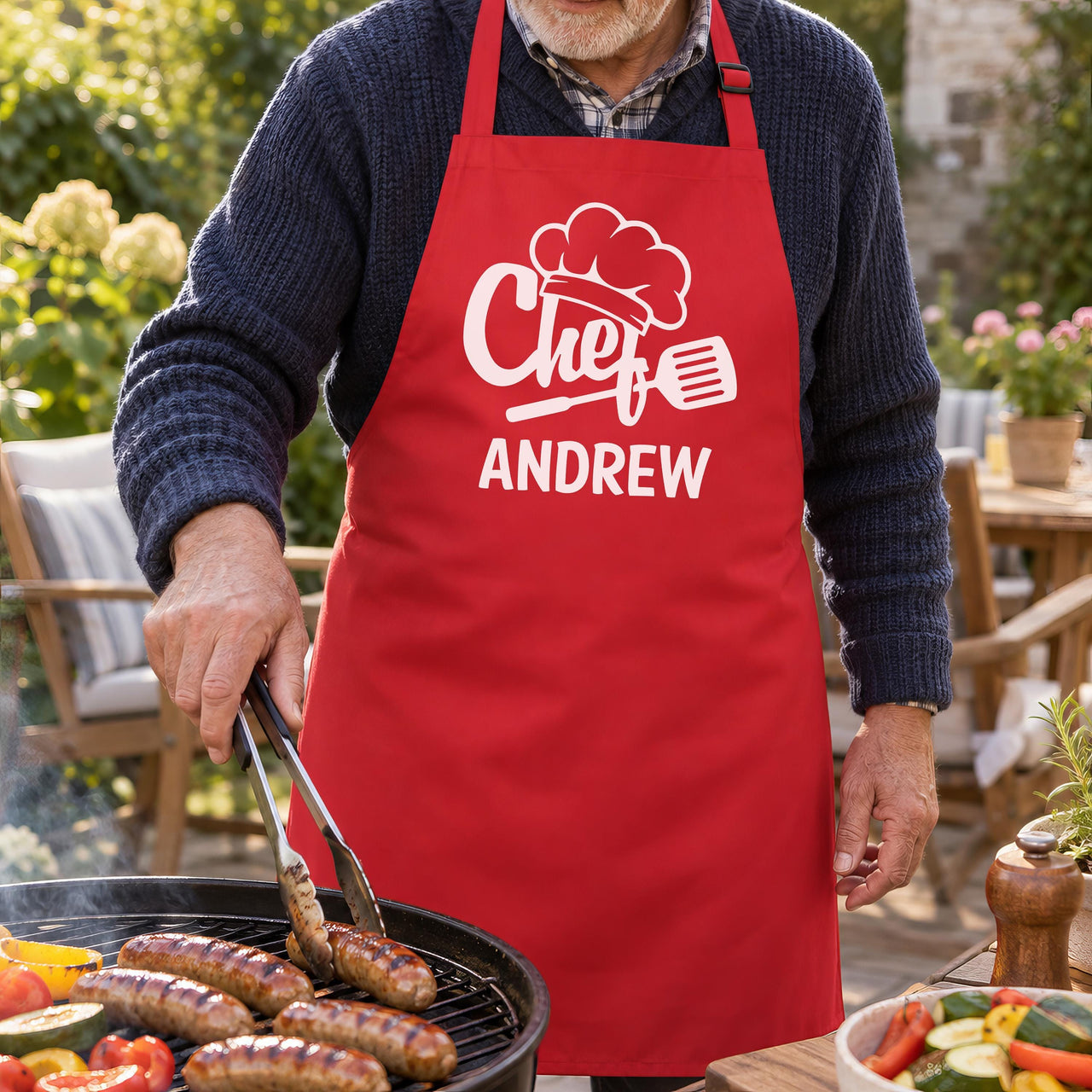 Red apron with white chef hat design and "ANDREW", worn while grilling sausages at a garden barbecue - personalised apron