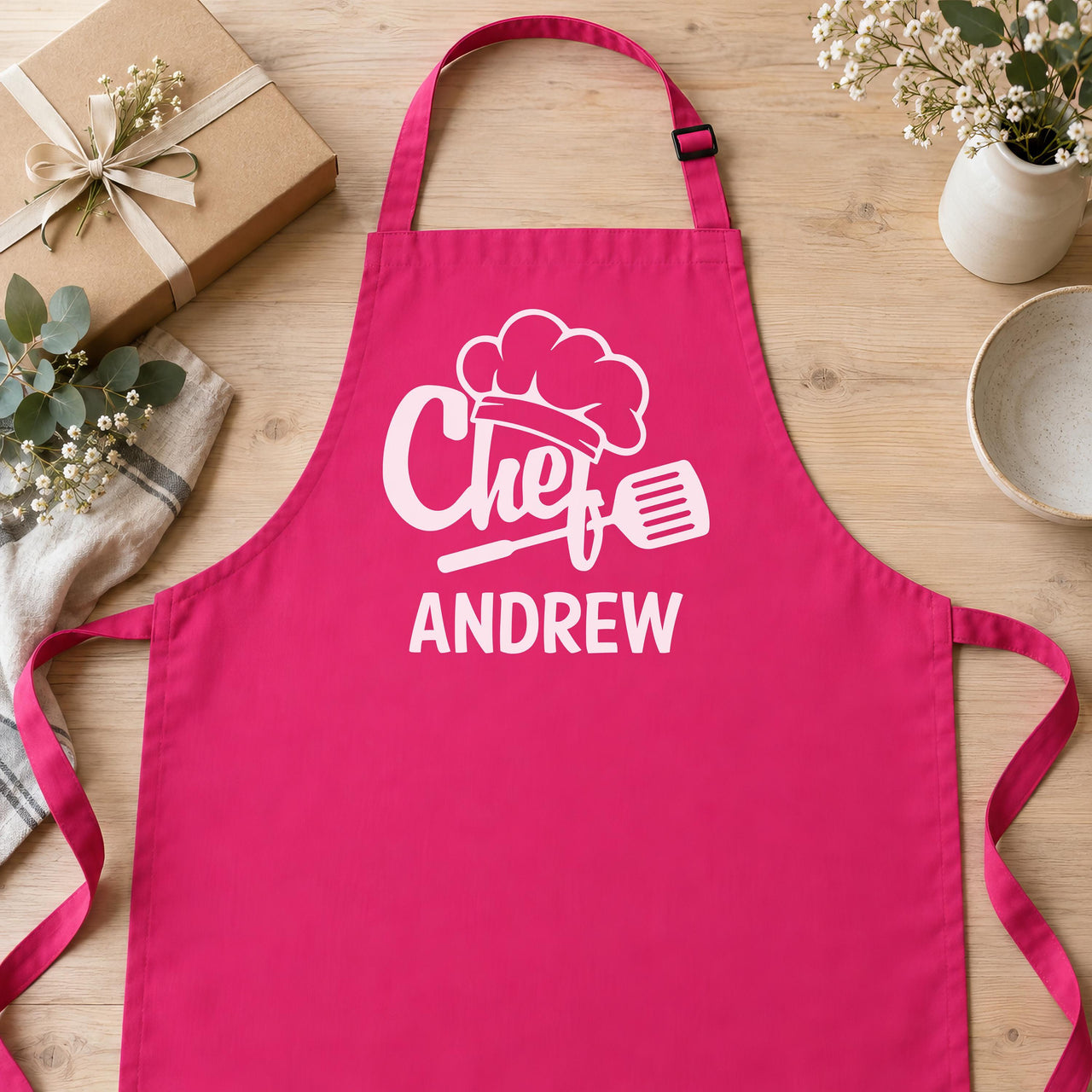 Pink apron laid flat with pale pink "Chef" hat, spatula and "ANDREW" print beside a wrapped gift - personalised apron