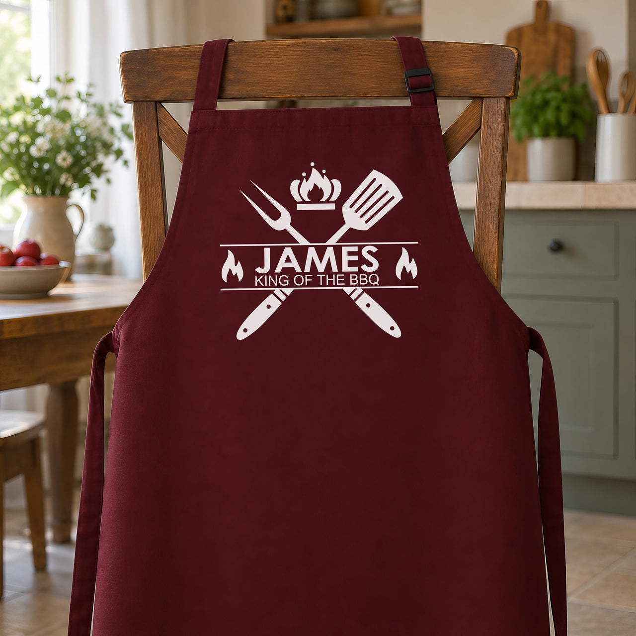 Burgundy apron with white "JAMES King of the BBQ" print hanging on a wooden kitchen chair - personalised apron