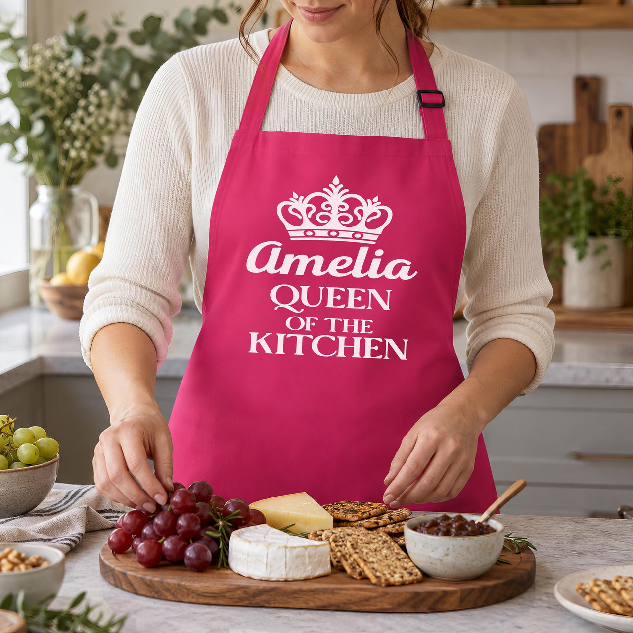 Pink apron with crown and 'Amelia QUEEN OF THE KITCHEN', arranging a cheese board - personalised apron