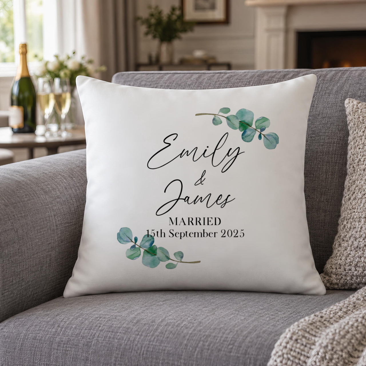 White wedding cushion with "Emily & James" and "Married" date on a grey sofa beside champagne glasses - personalised cushion