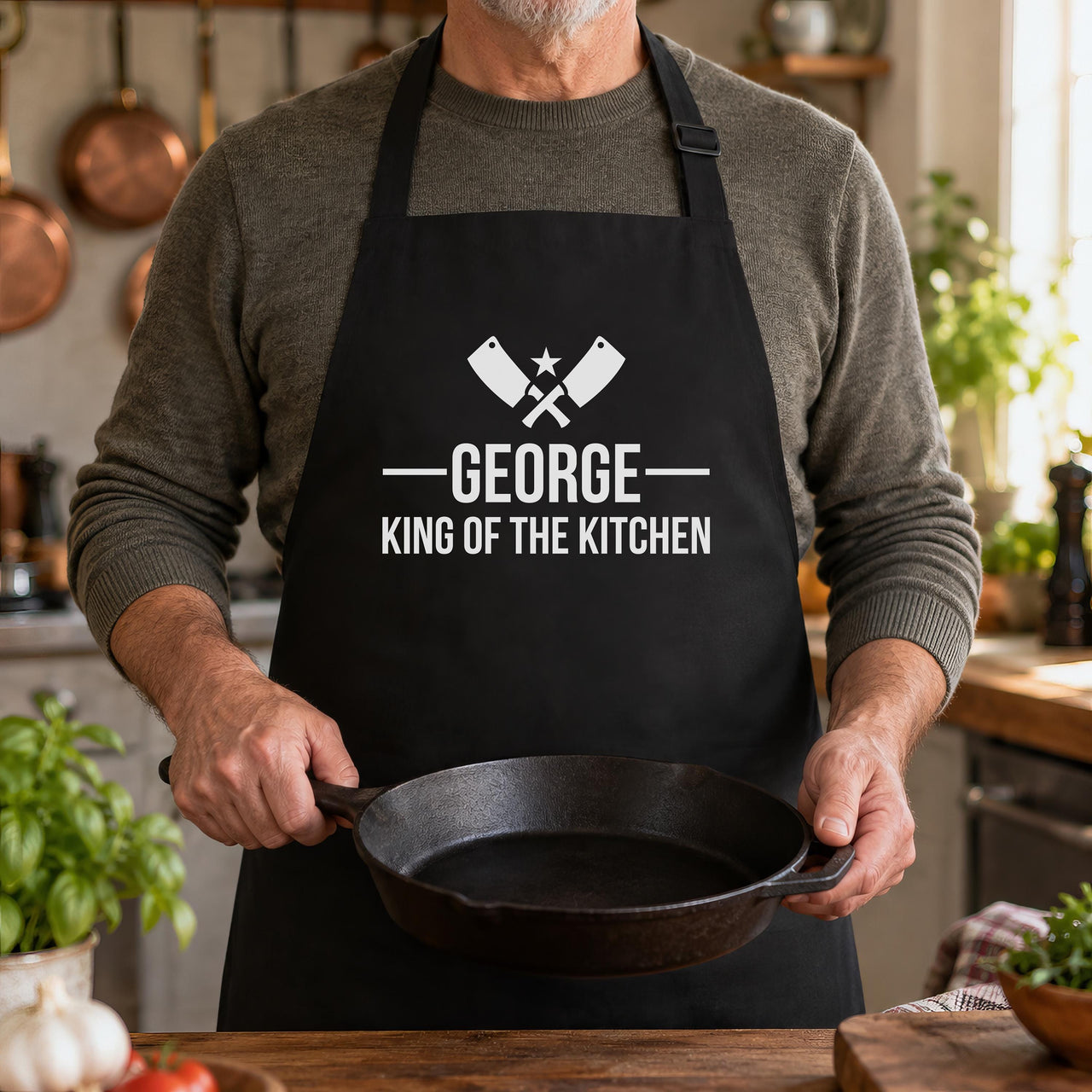 Man holding a cast iron pan in a black apron printed 'GEORGE King of the Kitchen' with crossed cleavers - personalised apron