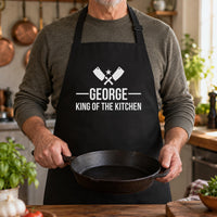 Man holding a cast iron pan in a black apron printed 'GEORGE King of the Kitchen' with crossed cleavers - personalised apron
