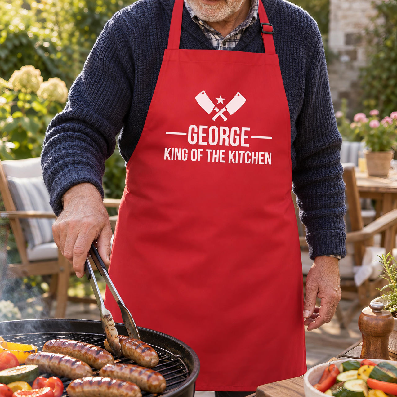 Man grilling sausages on a garden BBQ in a red apron with white 'GEORGE King of the Kitchen' text - personalised apron