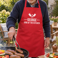 Man grilling sausages on a garden BBQ in a red apron with white 'GEORGE King of the Kitchen' text - personalised apron