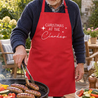 Man grilling sausages outdoors in a red apron printed 