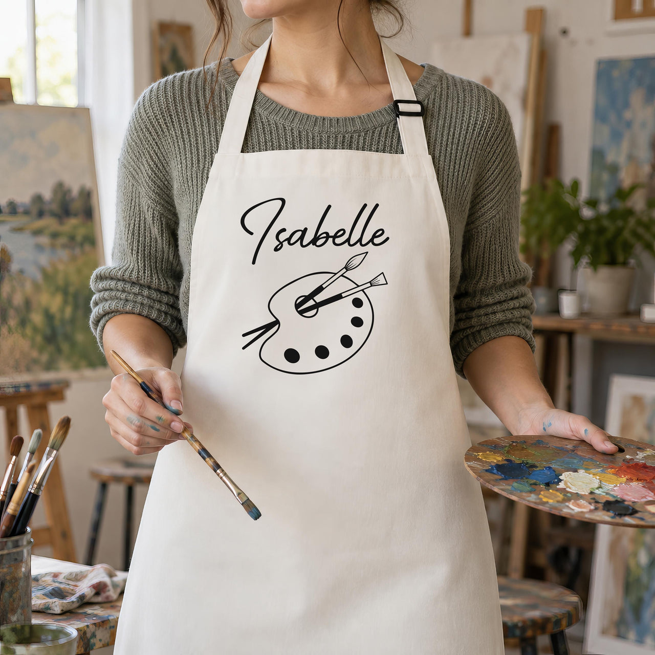 Natural apron with black 'Isabelle' and palette print, worn by an artist holding a brush - personalised apron