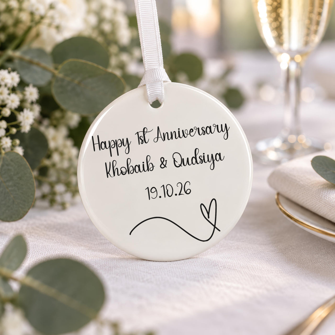 Anniversary ceramic bauble: script Happy 1st Anniversary Khobaib & Qudsiya 19.10.26, heart line, on a table with flowers