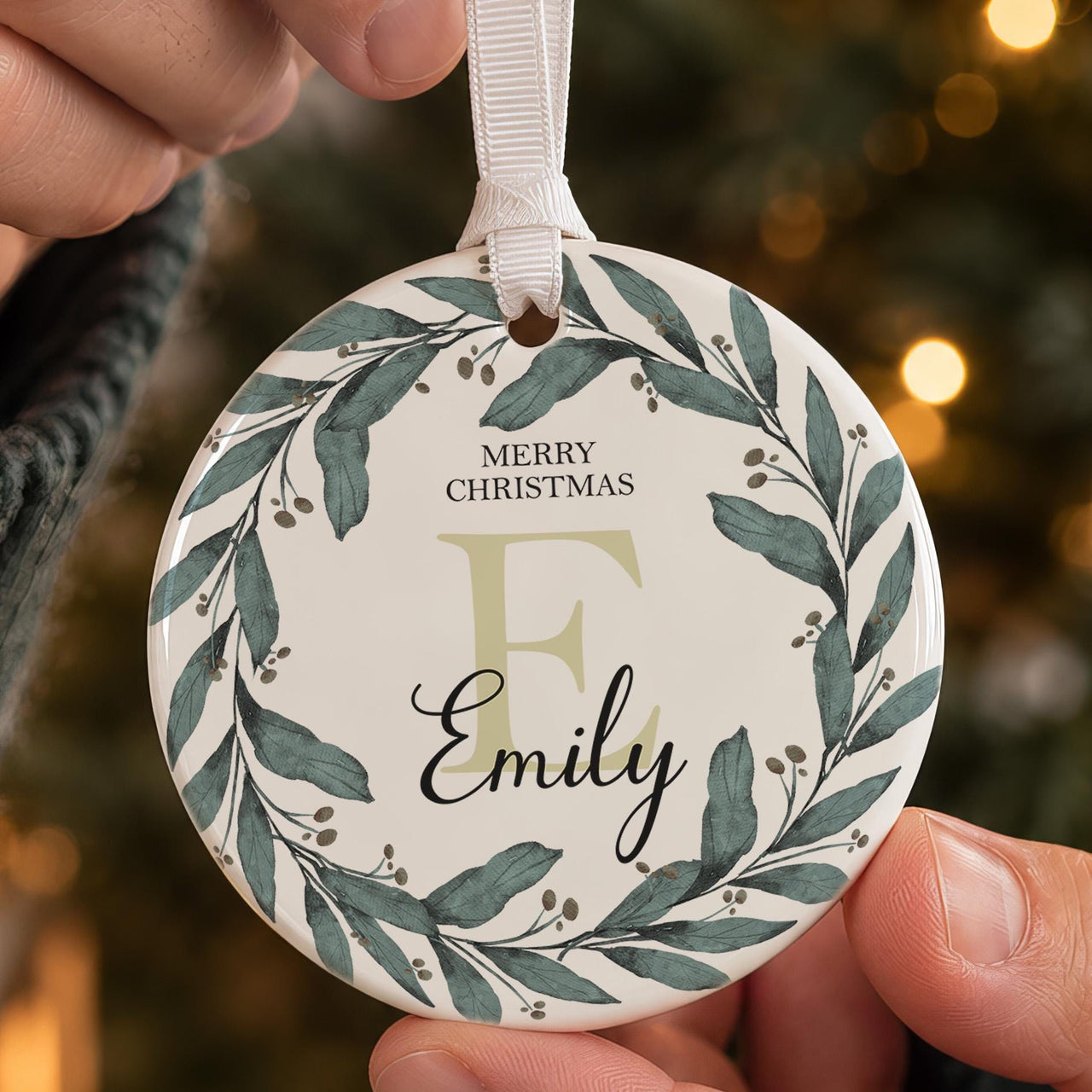 Wreath initial ceramic bauble held in front of a tree: dark green leaf wreath, Merry Christmas, pale E and Emily in script