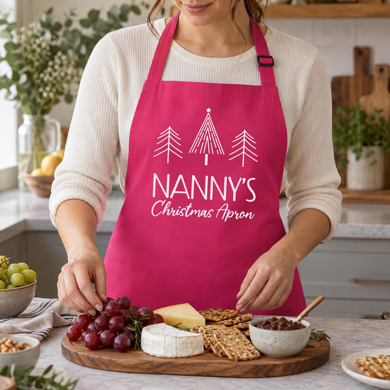 Pink Nanny's Christmas Apron with white tree print, worn while laying out a cheese board