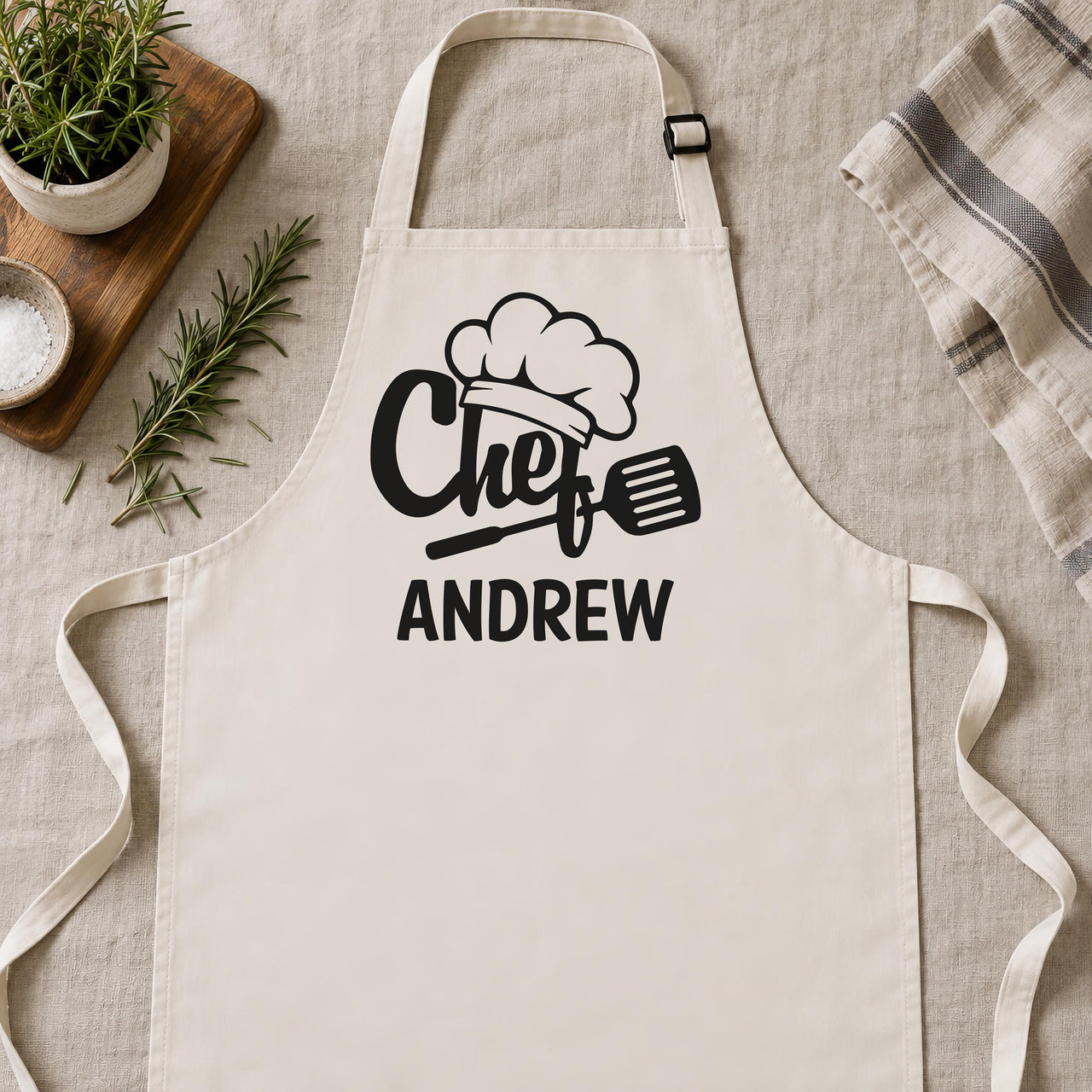 Natural cream apron laid flat with black "Chef" hat and spatula design and the name "ANDREW" - personalised apron