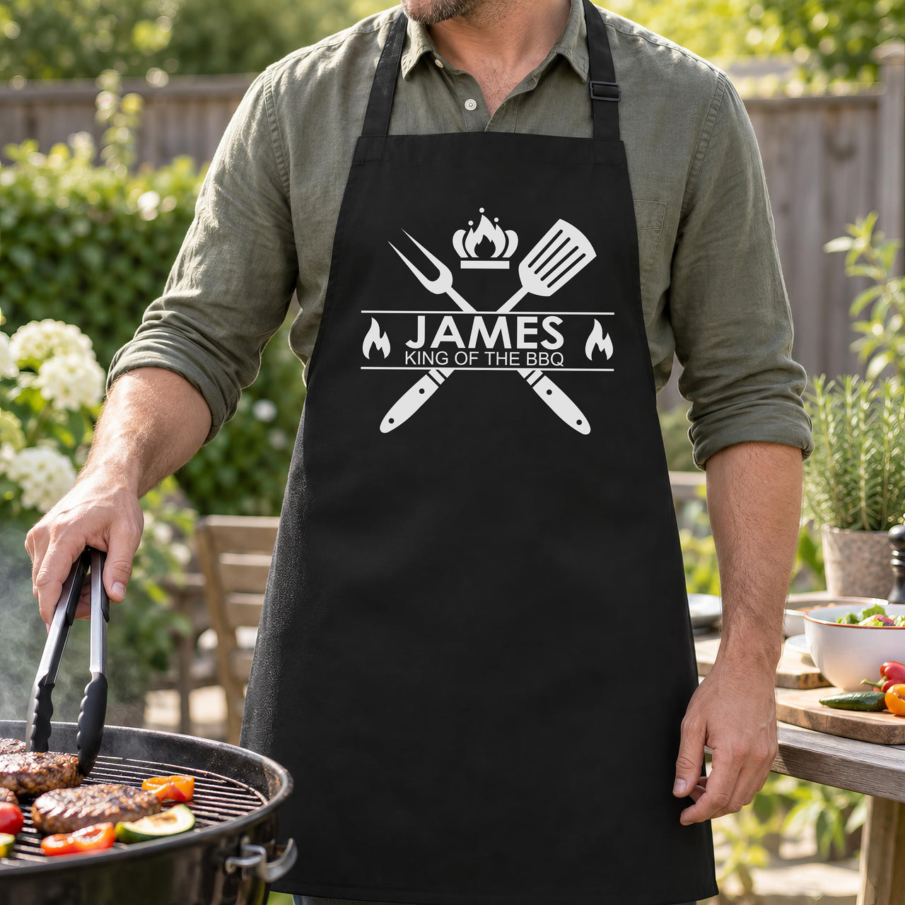 Black apron with white "JAMES King of the BBQ" design worn at a garden grill cooking burgers - personalised apron