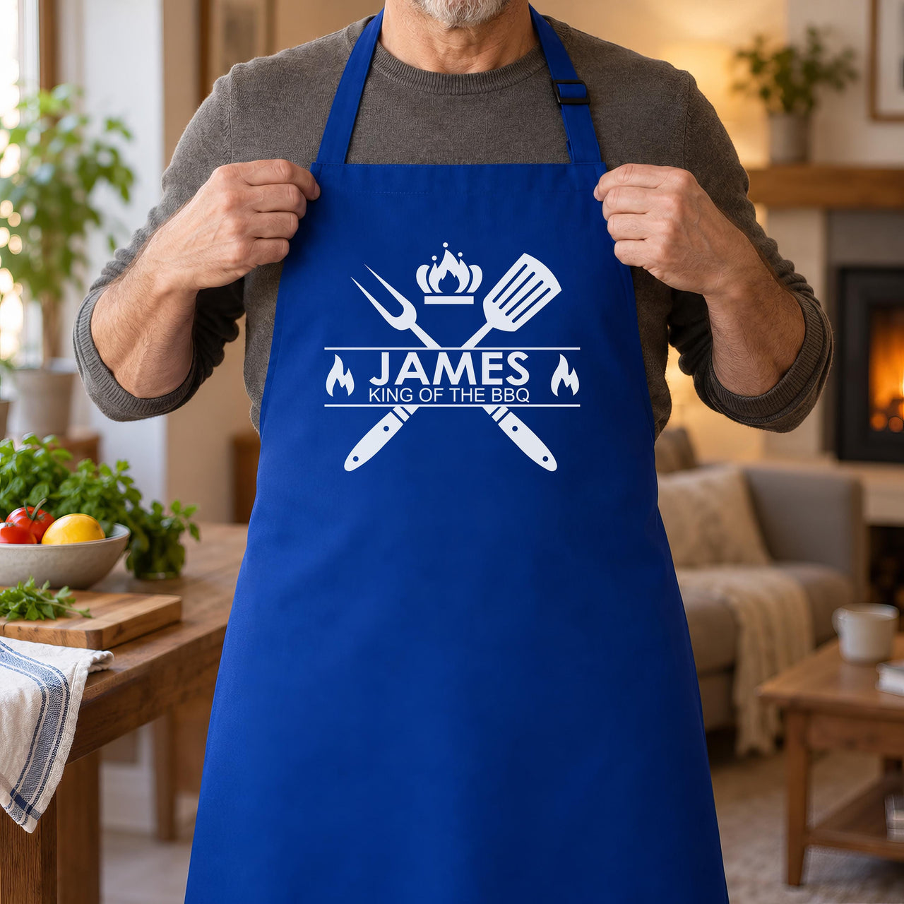 Man holding the straps of a royal blue apron printed "JAMES King of the BBQ" in a cosy living room - personalised apron