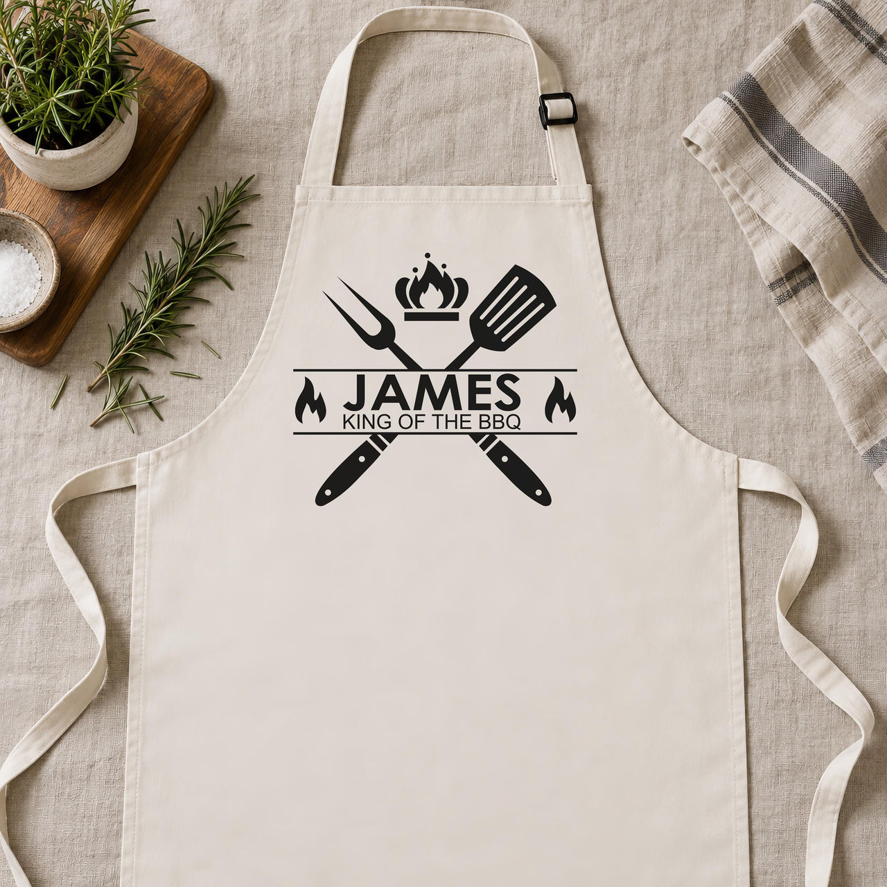 Natural cream apron laid flat with black "JAMES King of the BBQ" crest, fork, spatula and flames - personalised apron